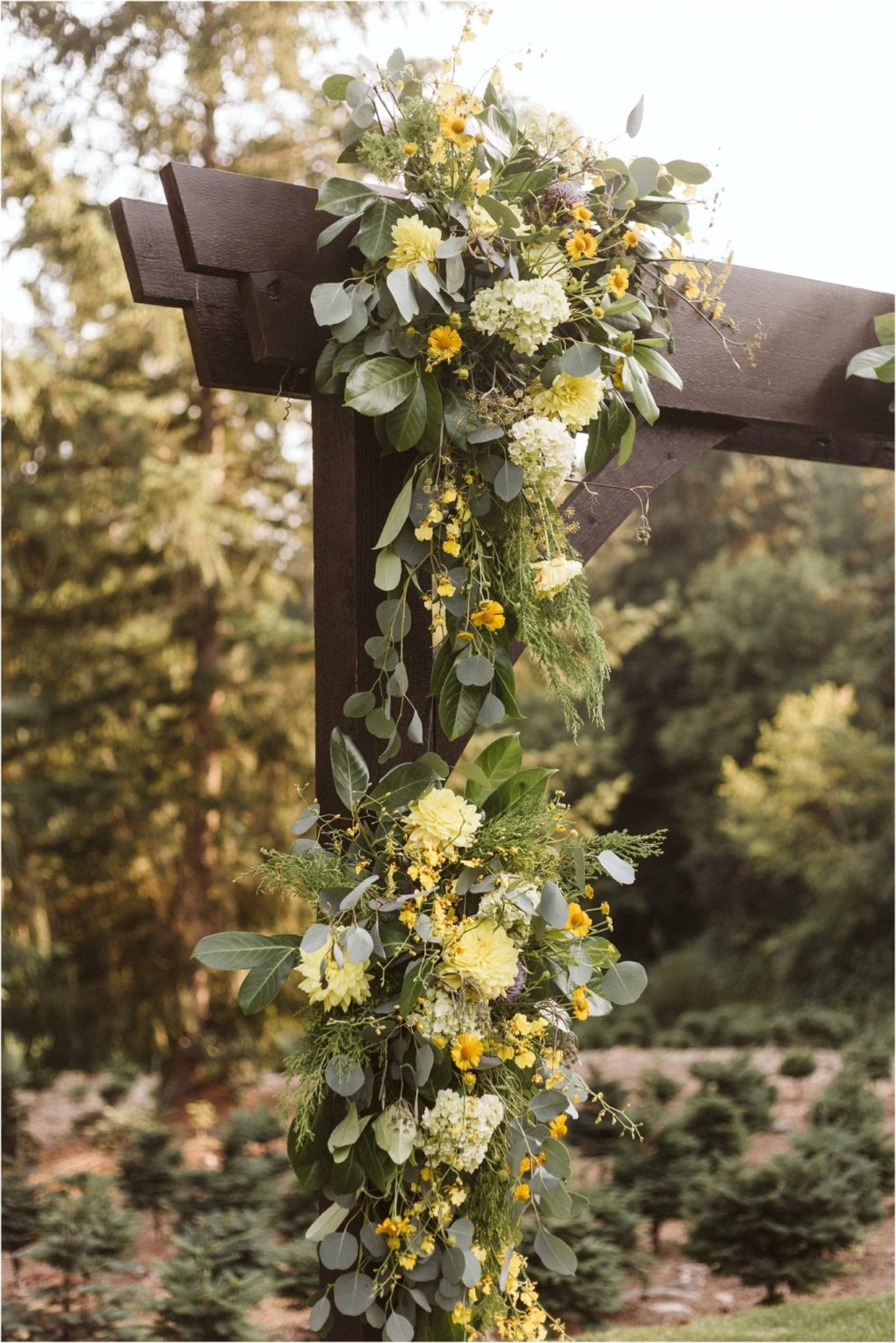 Premier snohomish & seattle wedding photographer | gsquared weddings photography a wooden arch decorated with lush greenery and clusters of yellow and white flowers perfect for a snohomish seattle wedding or engagement set outdoors with blurred trees in the background | serving snohomish seattle                   & beyond