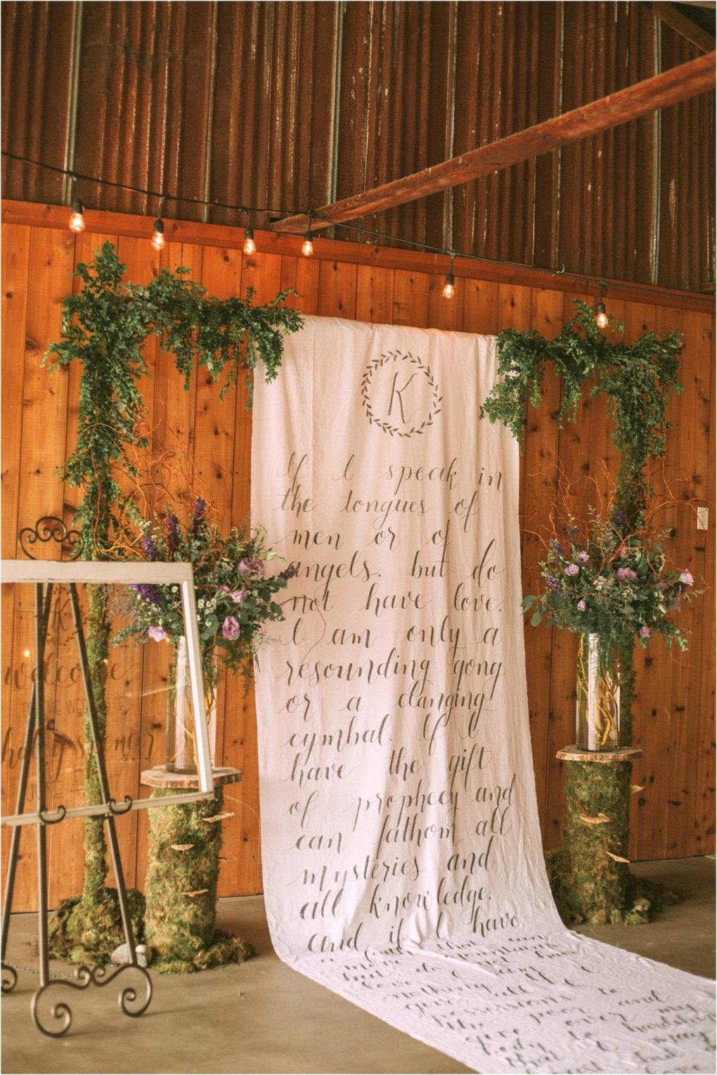 Premier snohomish & seattle wedding photographer | gsquared weddings photography a rustic indoor snohomish wedding backdrop with a white cloth featuring handwritten script flanked by lush flower arrangements on tree stumps greenery overhead and string lights against wooden walls An empty easel stands to the side | serving snohomish seattle                   & beyond
