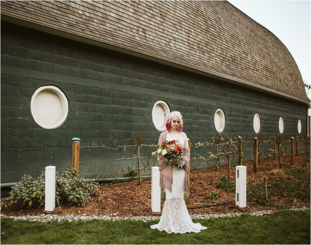 Premier snohomish & seattle wedding photographer | gsquared weddings photography a bride in a white lace gown and pink shawl holds a bouquet of flowers standing in front of a green building with round windowsa charming snohomish wedding scenesurrounded by a mulched garden | serving snohomish seattle                   & beyond