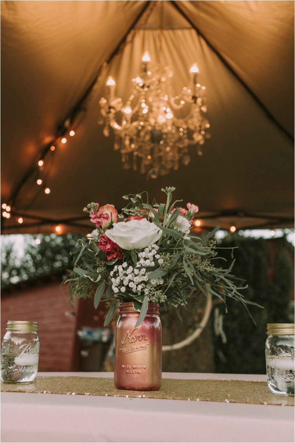 Premier snohomish & seattle wedding photographer | gsquared weddings photography a floral arrangement in a painted mason jar sits on a table under a lit chandelier with string lights above and two jars of water on either side creating a rustic romantic ambiance perfect for a snohomish or seattle wedding engagement | serving snohomish seattle                   & beyond