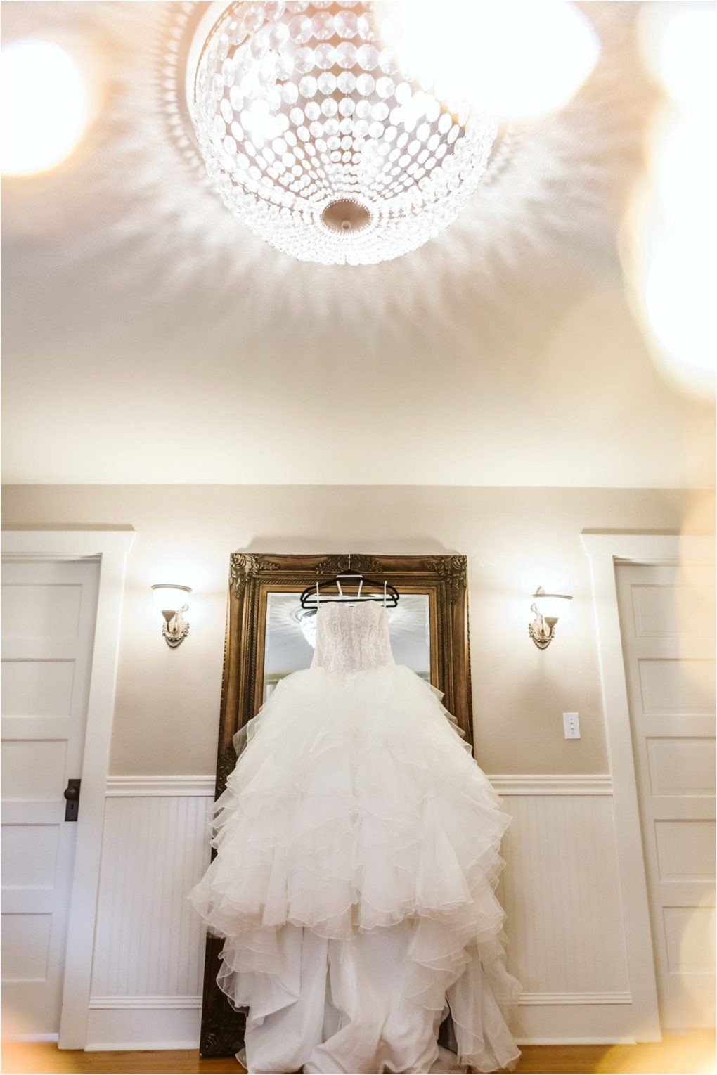 Premier snohomish & seattle wedding photographer | gsquared weddings photography a white ruffled wedding dress hangs on a mirror in an elegant room with white paneled wallsperfect for a snohomish seattle wedding or engagement complete with sparkling chandelier and graceful wall sconces | serving snohomish seattle                   & beyond
