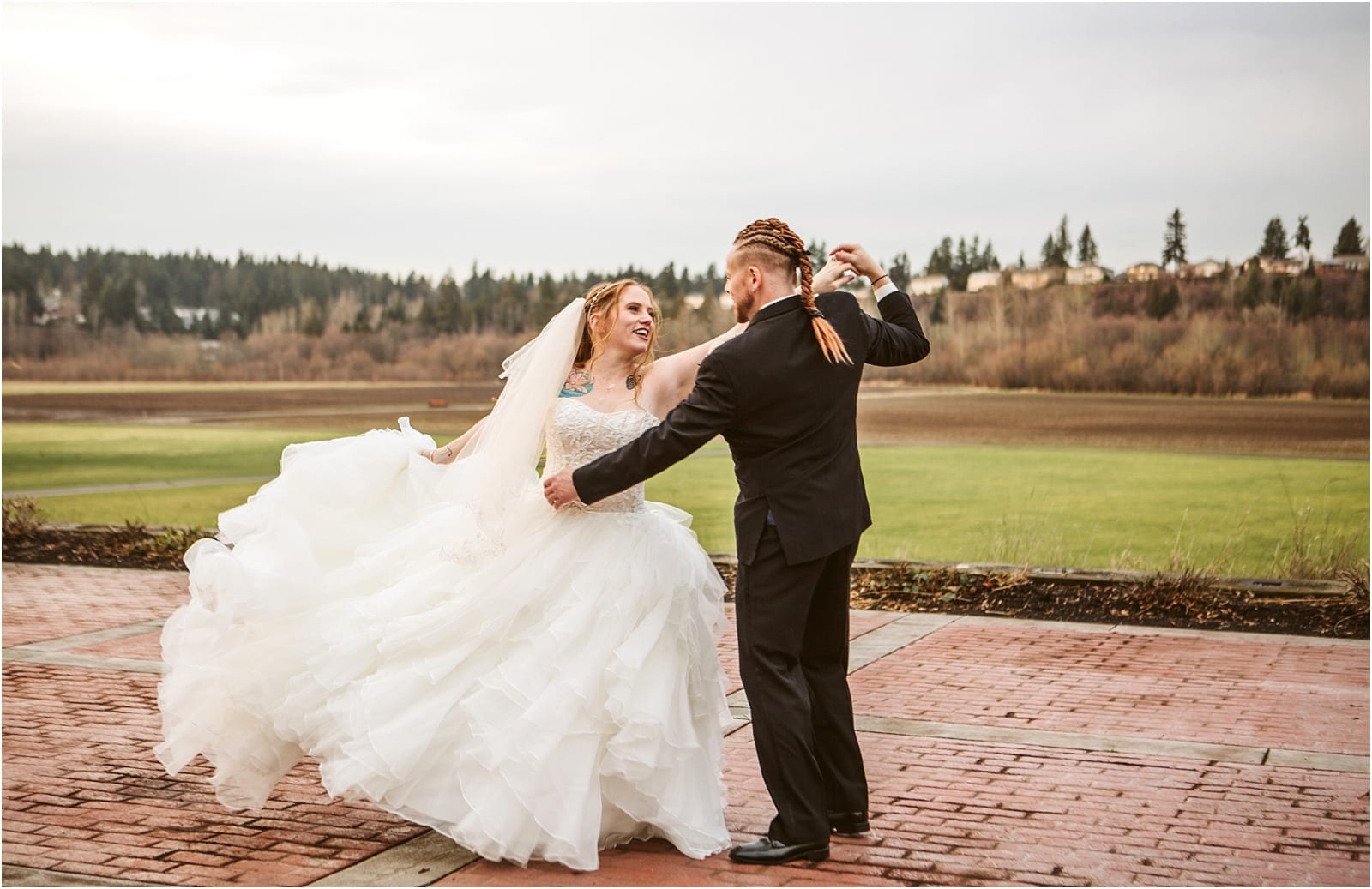 Premier Snohomish & Seattle Wedding Photographer | GSquared Weddings Photography A bride in a white gown and veil twirls joyfully with her groom in a black suit on a brick patio, capturing a magical Snohomish Seattle wedding moment amid grassy fields and trees under a cloudy sky.                                   | Serving Snohomish, Seattle ,                  & Beyond