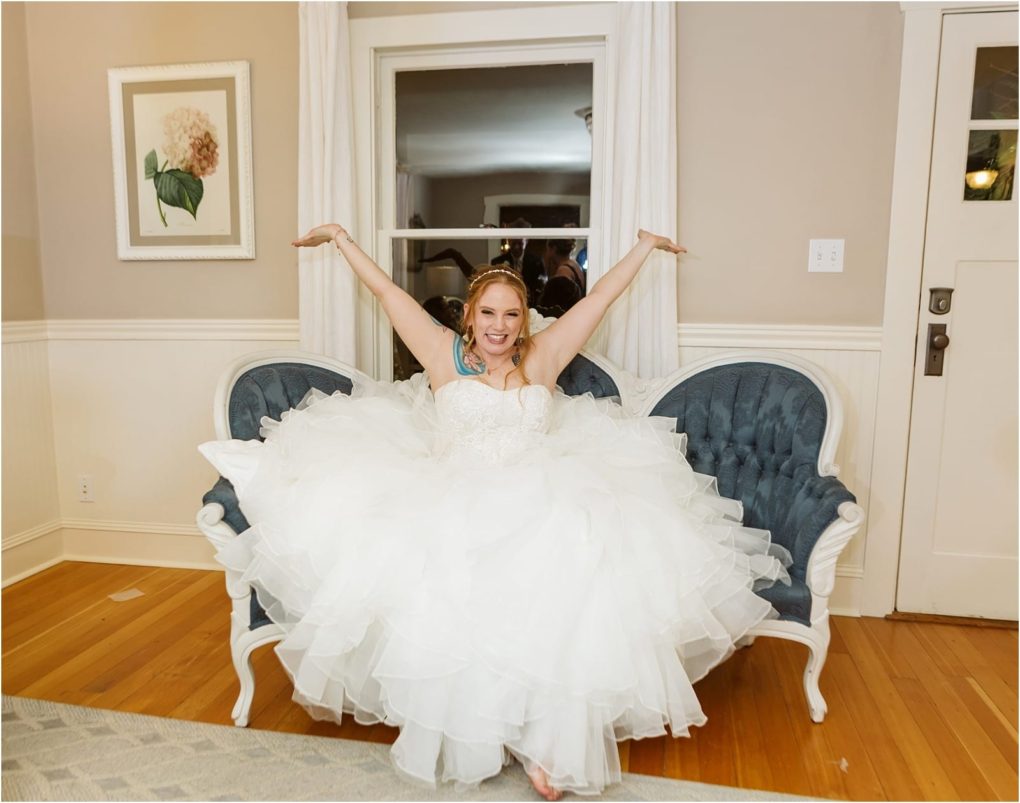 Premier snohomish & seattle wedding photographer | gsquared weddings photography a smiling bride in a white wedding dress sits on a blue loveseat with her arms raised joyfully Indoors at a snohomish seattle wedding a window and framed flower artwork are seen on the wall behind her | serving snohomish seattle                   & beyond