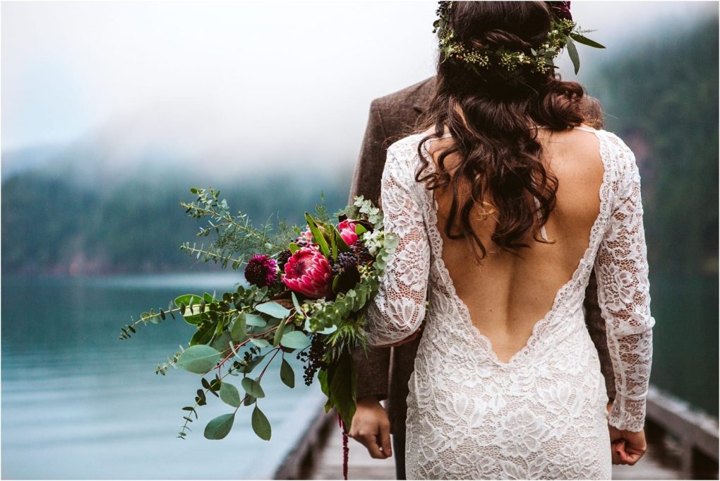 Premier snohomish & seattle wedding photographer | gsquared weddings photography a bride in a lace open back dress and flower crown stands holding a bouquet by a lake facing her partner Misty mountains and water create a serene backgroundperfect for a snohomish or seattle wedding engagement | serving snohomish seattle                   & beyond