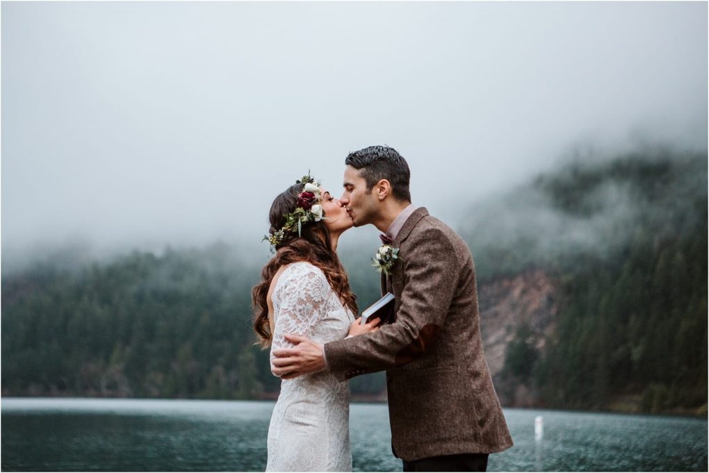 Premier snohomish & seattle wedding photographer | gsquared weddings photography a couple dressed in wedding attire shares a kiss by a misty lake near snohomish surrounded by forested mountains The bride wears a white lace dress and floral crown the groom in a brown suit holds a small book Perfect for a seattle wedding engagement | serving snohomish seattle                   & beyond