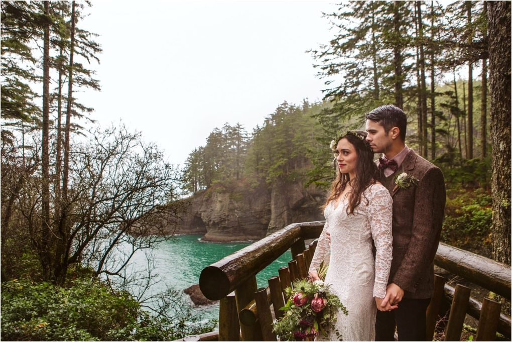 Premier snohomish & seattle wedding photographer | gsquared weddings photography a bride in a white lace dress and a groom in a brown suit stand on a wooden deck overlooking a forested coastal cliff and turquoise water holding hands and gazing into the distance at their snohomish seattle wedding | serving snohomish seattle                   & beyond