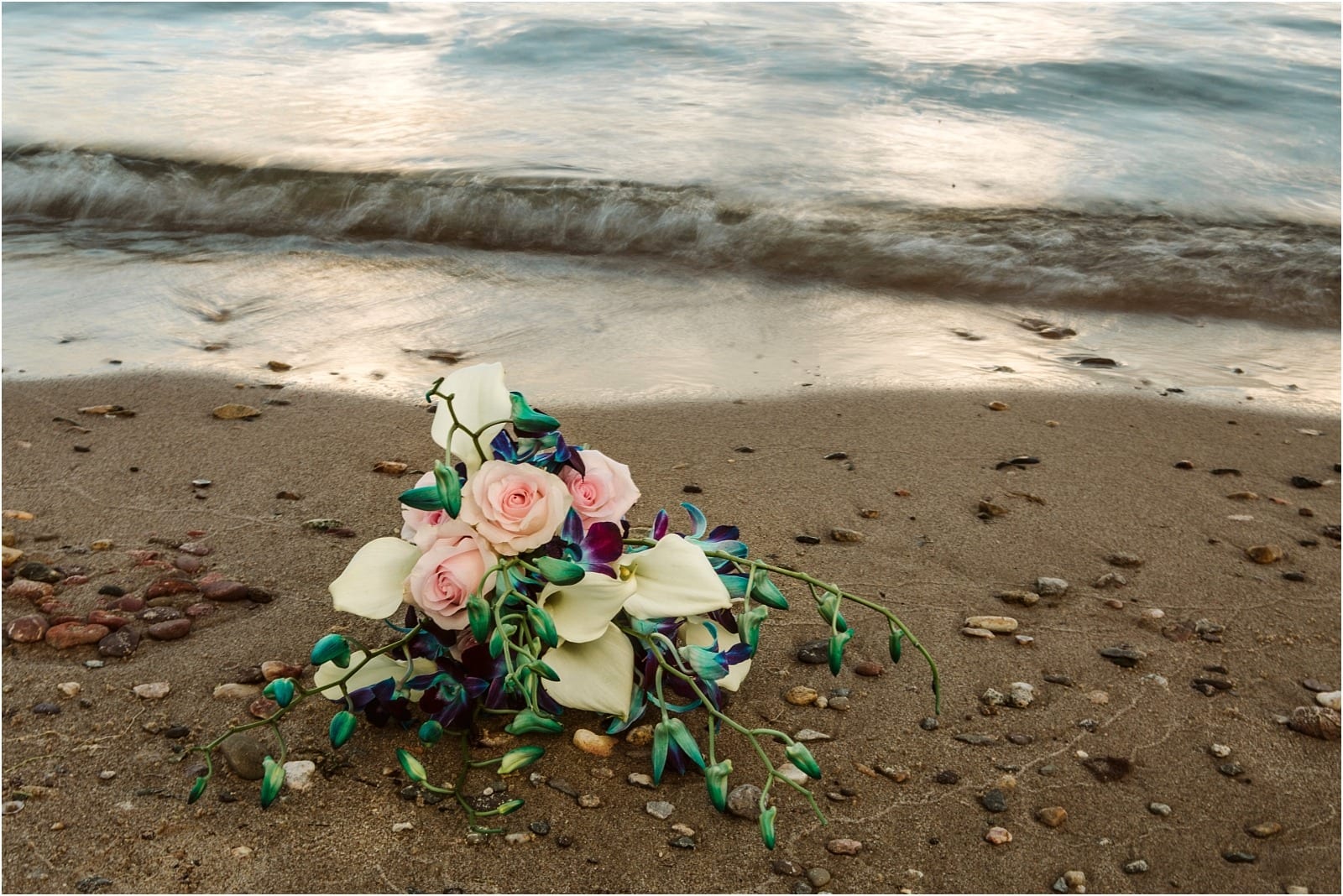 Premier Snohomish & Seattle Wedding Photographer | GSquared Weddings Photography A bouquet with pink roses, white calla lilies, and blue-purple flowers lies on a sandy beach near gentle waves—perfect for a Snohomish or Seattle wedding engagement, surrounded by small pebbles.                                   | Serving Snohomish, Seattle ,                  & Beyond