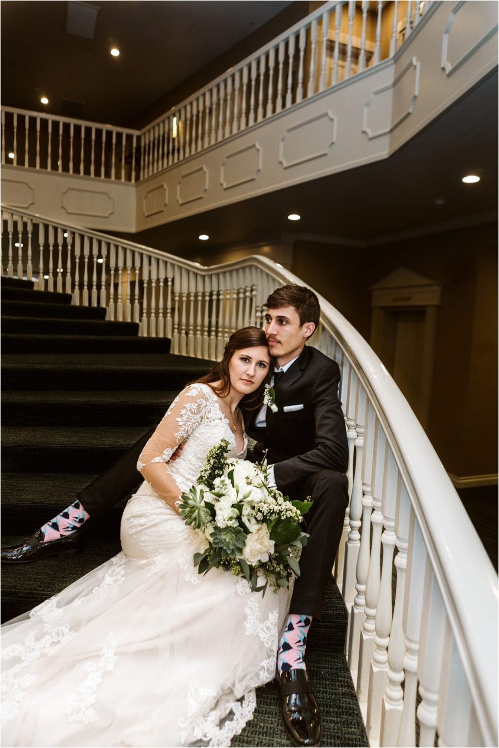 Premier snohomish & seattle wedding photographer | gsquared weddings photography a bride in a lace wedding dress and a groom in a dark suit with colorful socks sit on a curved staircase during their snohomish seattle wedding the bride holding white flowers as they look thoughtfully into the distance | serving snohomish seattle                   & beyond