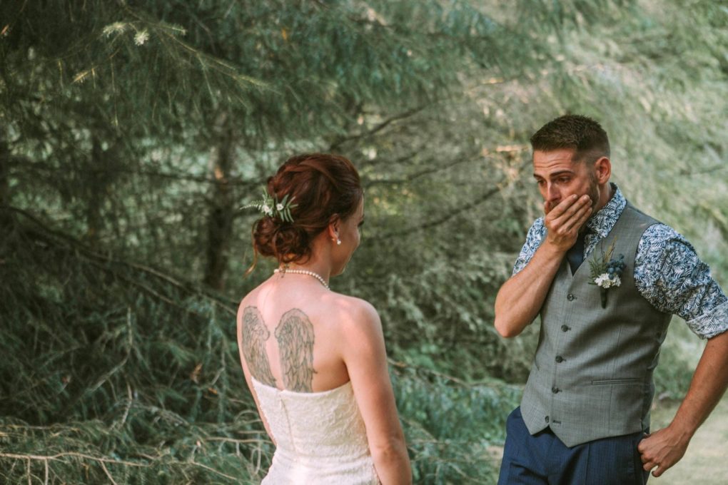 Premier snohomish & seattle wedding photographer | gsquared weddings photography a bride in a white dress with angel wing tattoos stands facing her groom in a forest near snohomish he covers his mouth in joyful surprise capturing an unforgettable moment during their seattle wedding engagement | serving snohomish seattle & beyond Premier snohomish & seattle wedding photographer | gsquared weddings photography a bride in a white dress with angel wing tattoos stands facing her groom in a forest near snohomish he covers his mouth in joyful surprise capturing an unforgettable moment during their seattle wedding engagement | serving snohomish seattle & beyond