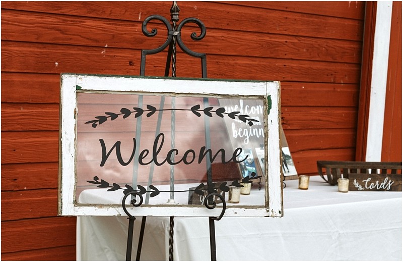 Premier snohomish & seattle wedding photographer | gsquared weddings photography a glass sign with black lettering that reads welcome and leafy designs is displayed on a stand in front of a red wooden wall at a snohomish seattle wedding beside a white table with candles and a basket labeled cards  | serving snohomish seattle                   & beyond