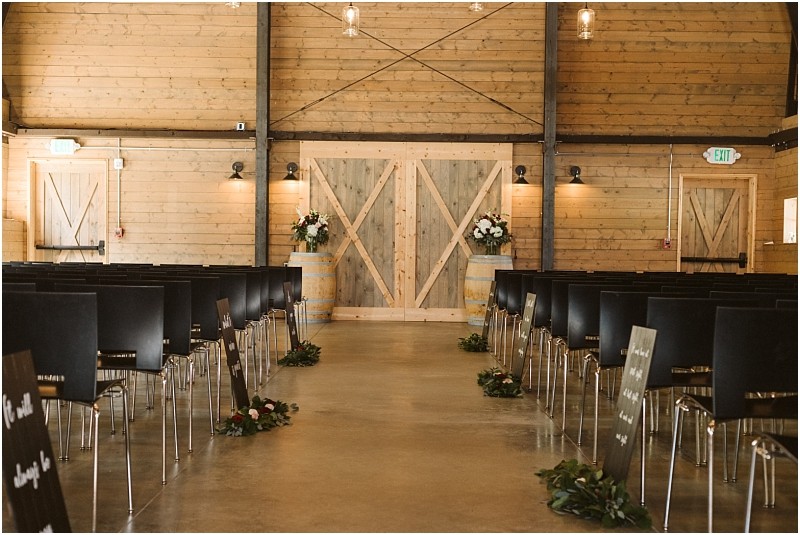 Premier snohomish & seattle wedding photographer | gsquared weddings photography a rustic indoor snohomish wedding ceremony setup features rows of black chairs wooden walls and large barn doors at the front Floral arrangements and greenery line the aisle with black signs with white writing standing along the aisle | serving snohomish seattle                   & beyond