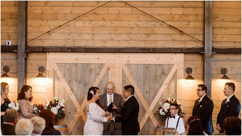 Premier snohomish & seattle wedding photographer | gsquared weddings photography a bride and groom stand facing each other holding hands at an indoor snohomish wedding ceremony surrounded by their wedding party and guests with a wooden barn door and floral arrangements in the background | serving snohomish seattle                   & beyond