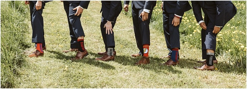 Premier snohomish & seattle wedding photographer | gsquared weddings photography five men in suits at a snohomish wedding lift their pant legs to reveal colorful mismatched novelty socks with various patterns and words standing in a row on a grassy path surrounded by green grass and yellow flowers | serving snohomish seattle                   & beyond