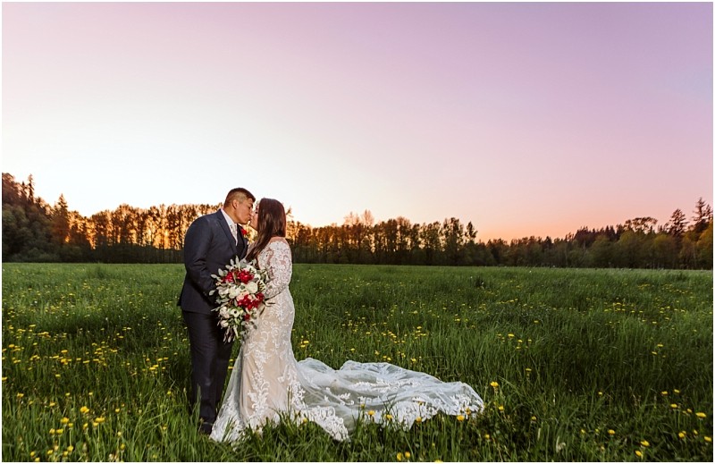 Premier snohomish & seattle wedding photographer | gsquared weddings photography a bride and groom stand in a green field at sunset kissing during their snohomish wedding The bride wears a long white lace dress with a train and holds a bouquet of red and white flowers Trees line the horizon | serving snohomish seattle                   & beyond