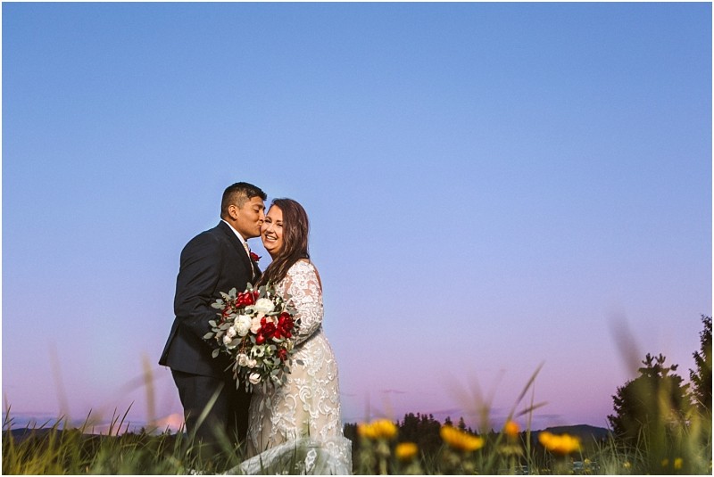 Premier snohomish & seattle wedding photographer | gsquared weddings photography a groom in a dark suit kisses his smiling bride who holds a large bouquet of red and white flowers They stand in tall grass dotted with yellow blooms as the sun sets capturing a magical snohomish wedding under a clear purple and blue sky | serving snohomish seattle                   & beyond