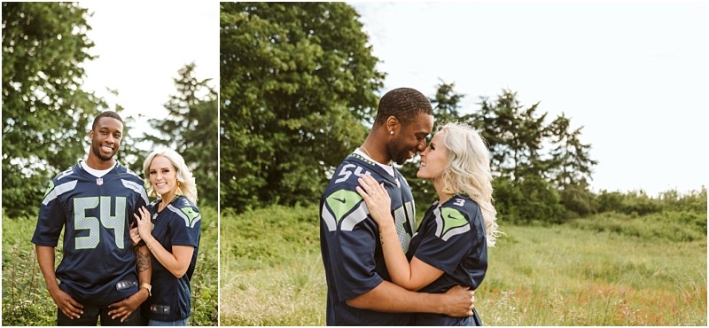 Premier snohomish & seattle wedding photographer | gsquared weddings photography a couple wearing matching dark blue football jerseys stands close together outdoors smiling and embracing in a grassy field with trees in the background during their snohomish seattle engagement | serving snohomish seattle                   & beyond