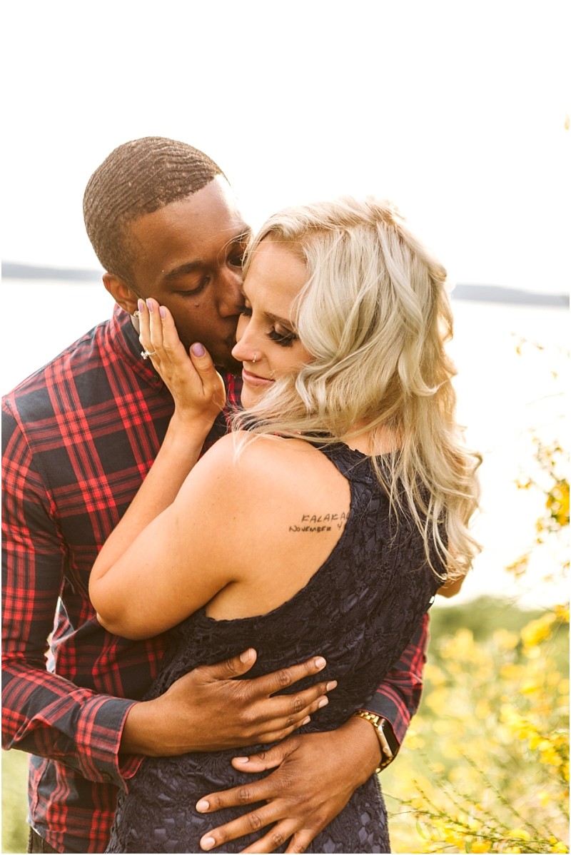 Premier snohomish & seattle wedding photographer | gsquared weddings photography a man in a red plaid shirt embraces and kisses a smiling woman with blonde hair in a dark dress outdoors with sunlight and greenerycapturing a romantic snohomish seattle engagement moment | serving snohomish seattle                   & beyond