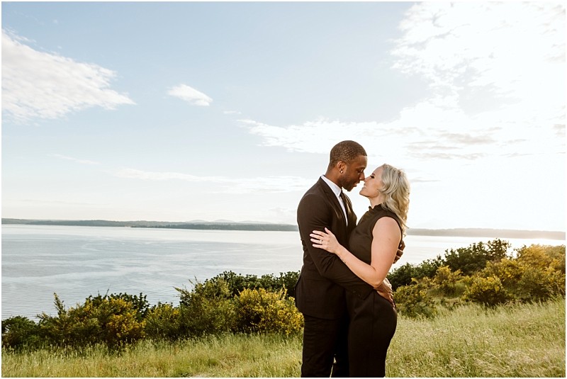 Premier snohomish & seattle wedding photographer | gsquared weddings photography a couple dressed in formal black attire stands closely together on a grassy hill overlooking a large body of water under a partly cloudy sky embracing and gazing into each others eyes during their snohomish wedding engagement | serving snohomish seattle                   & beyond