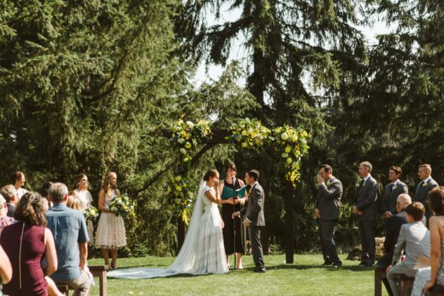 Premier snohomish & seattle wedding photographer | gsquared weddings photography a bride and groom stand under a floral arch exchanging vows outdoors at a snohomish seattle wedding surrounded by bridesmaids groomsmen and seated guests with tall pine trees in the background | serving snohomish seattle                   & beyond