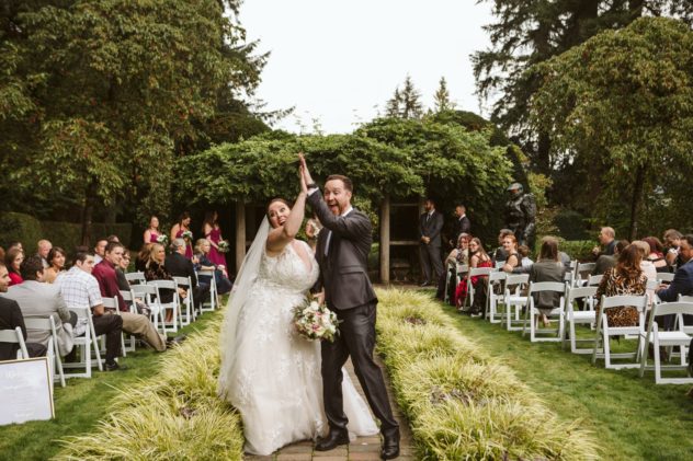 Premier snohomish & seattle wedding photographer | gsquared weddings photography a joyful bride and groom celebrate after their outdoor snohomish wedding ceremony holding hands and raising them in the air as guests seated on white chairs watch and smile Lush greenery surrounds the garden venue | serving snohomish seattle                   & beyond