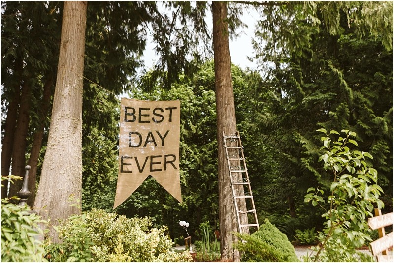 Premier snohomish & seattle wedding photographer | gsquared weddings photography a large fabric banner reading best day ever hangs between two tall trees in a lush green outdoor settinga perfect spot for a snohomish seattle wedding or engagement A wooden ladder leans against one tree with shrubs and plants filling the foreground | serving snohomish seattle                   & beyond