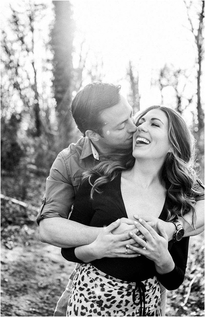 Premier snohomish & seattle wedding photographer | gsquared weddings photography a man embraces a smiling woman from behind playfully kissing her cheek as they stand on a sunlit forest path Both appear happy and relaxed with trees and sunlight in the backgroundcapturing a joyful snohomish engagement in black and white | serving snohomish seattle                   & beyond