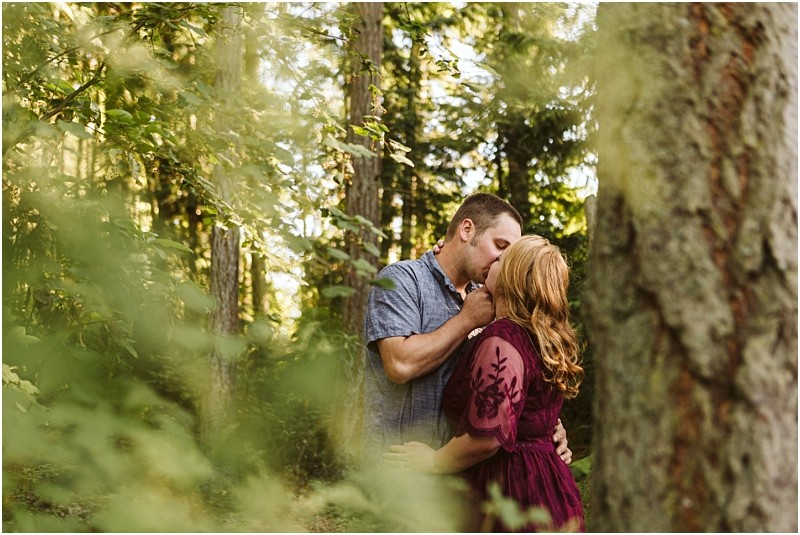 Premier snohomish & seattle wedding photographer | gsquared weddings photography a couple kisses in a sunlit forest near snohomish surrounded by green trees and foliage The man wears a blue shirt and the woman has long red hair and a burgundy dressan intimate natural seattle engagement moment | serving snohomish seattle                   & beyond