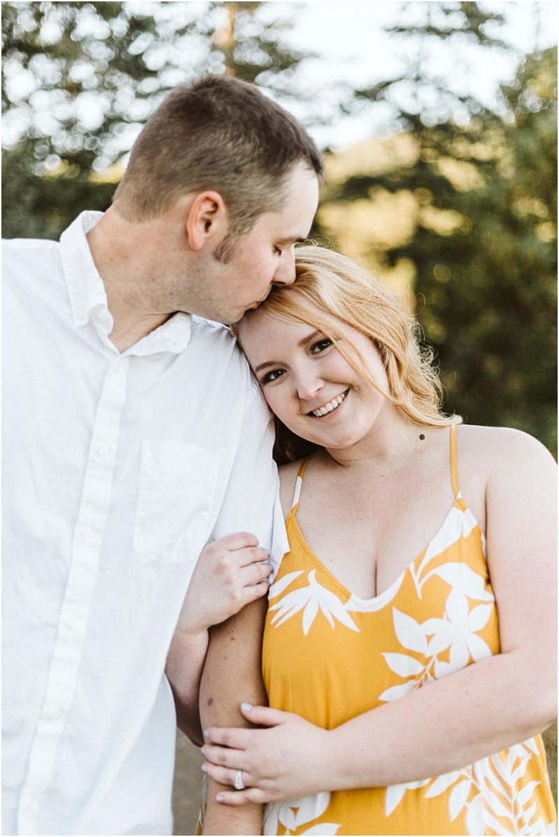 Premier snohomish & seattle wedding photographer | gsquared weddings photography a man in a white shirt kisses the forehead of a smiling woman in a yellow floral dress during their snohomish engagement The woman holds his arm as they stand outdoors surrounded by trees | serving snohomish seattle                   & beyond