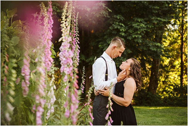 Premier Snohomish & Seattle Wedding Photographer | GSquared Weddings Photography A couple embraces lovingly in a sunlit garden, surrounded by tall pink flowers and lush greenery during their Snohomish Seattle wedding engagement. The man wears a white shirt and suspenders, while the woman gazes up at him affectionately in a black dress.                                   | Serving Snohomish, Seattle ,                  & Beyond