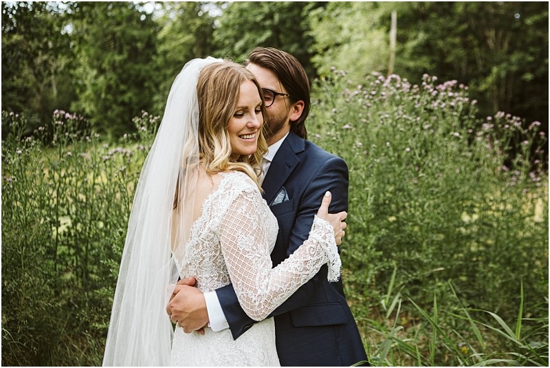 Premier snohomish & seattle wedding photographer | gsquared weddings photography a bride in a lace dress and veil smiles while being hugged by a groom in a blue suit and glasses standing close together among tall grass and wildflowers during their snohomish seattle wedding | serving snohomish seattle                   & beyond