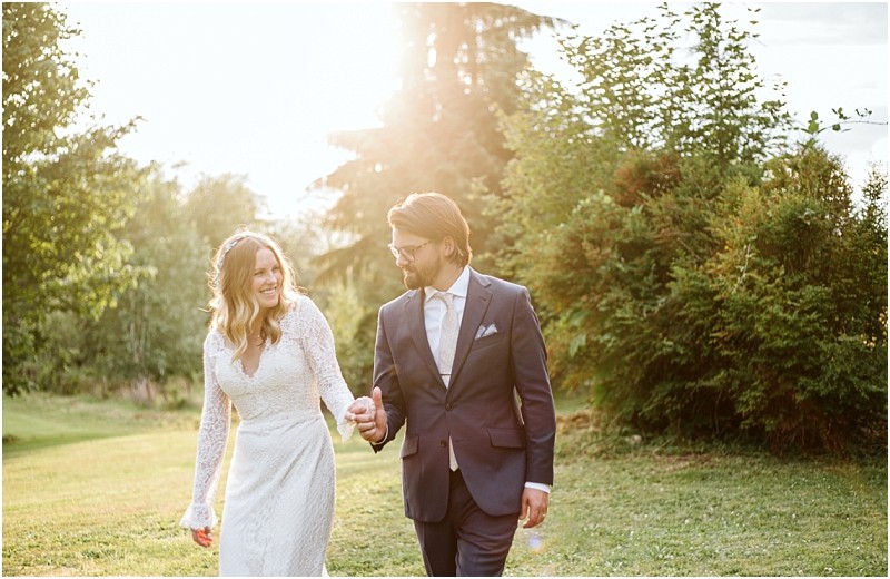 Premier snohomish & seattle wedding photographer | gsquared weddings photography a bride in a white lace dress and a groom in a navy suit walk hand in hand on a grassy lawn smiling at each other with sunlight streaming through the trees behind them during their snohomish seattle wedding | serving snohomish seattle                   & beyond