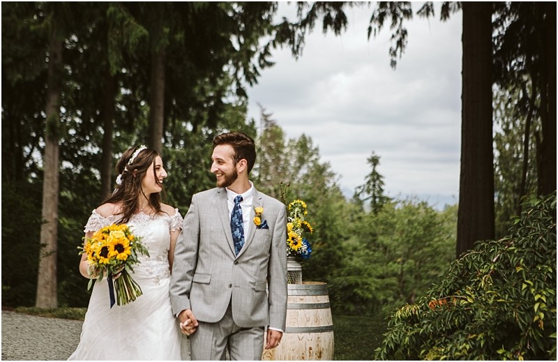 Premier snohomish & seattle wedding photographer | gsquared weddings photography a bride in a white dress holding sunflowers and a groom in a light gray suit smile at each other while walking outdoors at their snohomish seattle wedding surrounded by trees greenery and floral decorations on wooden barrels | serving snohomish seattle                   & beyond