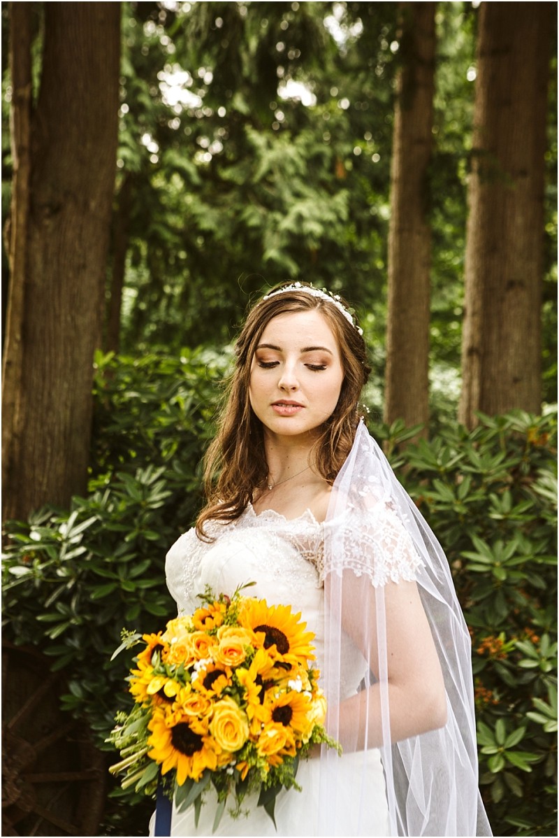 Premier snohomish & seattle wedding photographer | gsquared weddings photography a bride in a lace wedding dress and veil holds a bouquet of sunflowers and yellow roses standing outdoors in a wooded area with lush green foliagea perfect snohomish seattle wedding scene | serving snohomish seattle                   & beyond