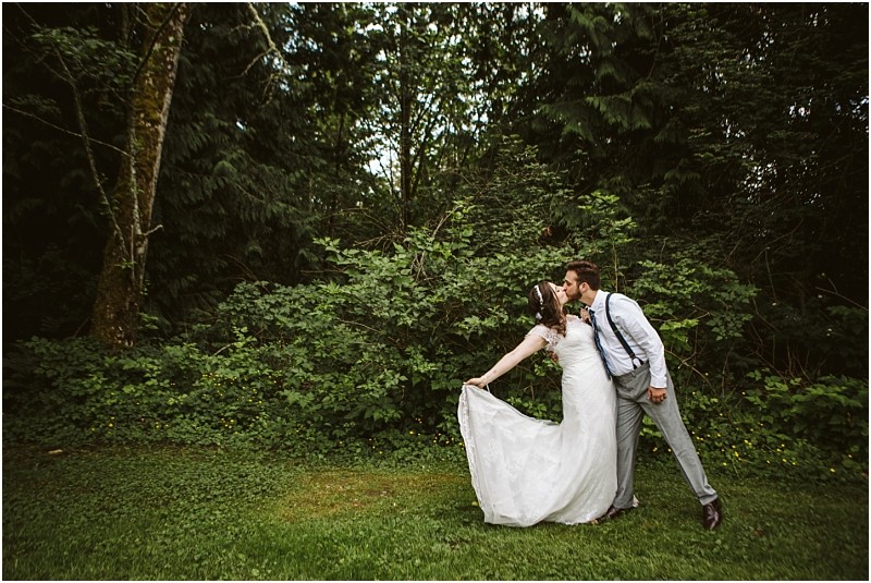 Premier snohomish & seattle wedding photographer | gsquared weddings photography a bride and groom stand on grass in front of dense green trees during their playful and romantic snohomish wedding the groom leans in to kiss the bride while she holds her dress and leans back both smiling joyfully | serving snohomish seattle                   & beyond
