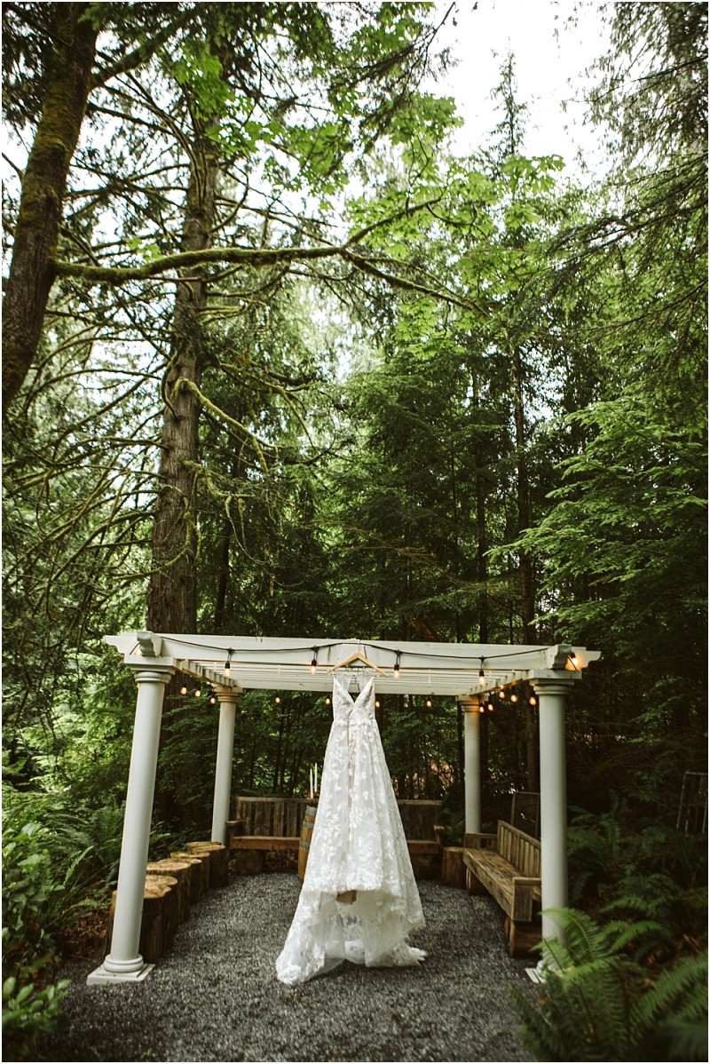 Premier snohomish & seattle wedding photographer | gsquared weddings photography a white lace wedding dress hangs from a pergola in a snohomish forest setting surrounded by tall trees and string lights with wooden benches arranged underneathperfect for a dreamy seattle wedding or engagement | serving snohomish seattle                   & beyond