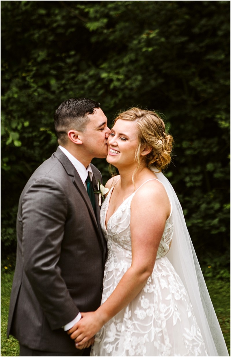 Premier snohomish & seattle wedding photographer | gsquared weddings photography a groom in a gray suit kisses the smiling bride on the cheek as they hold hands outdoors surrounded by lush greenery during their snohomish seattle wedding The bride wears a white lace dress and a veil | serving snohomish seattle                   & beyond