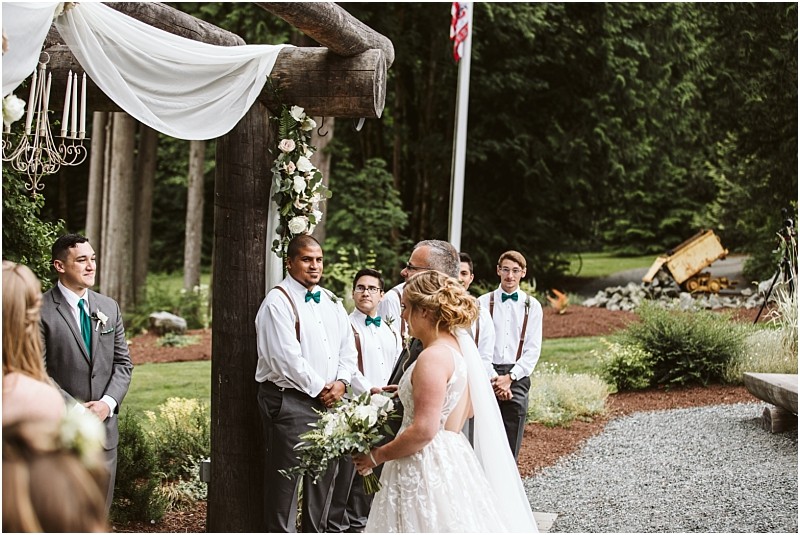 Premier snohomish & seattle wedding photographer | gsquared weddings photography a bride and groom stand together at an outdoor snohomish wedding ceremony facing an officiant under a wooden arch decorated with white fabric and flowers Groomsmen in white shirts and green bow ties look on surrounded by lush seattle greenery | serving snohomish seattle                   & beyond