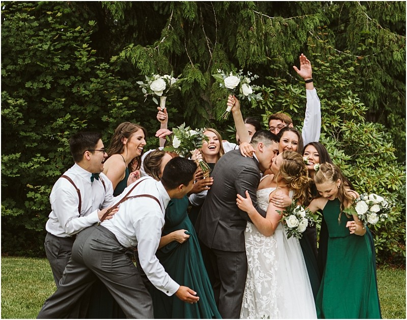 Premier snohomish & seattle wedding photographer | gsquared weddings photography a bride and groom kiss outdoors at their snohomish seattle wedding surrounded by a joyful party in green dresses and suspenders Bridesmaids cheer with bouquets groomsmen laugh and lush greenery forms the perfect backdrop for this magical engagement | serving snohomish seattle                   & beyond