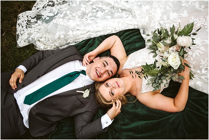 Premier snohomish & seattle wedding photographer | gsquared weddings photography a bride and groom lie on a green blanket smiling up at the camera during their snohomish wedding The bride holds a bouquet of white flowers while the grooms gray suit and green tie complement her lacy dress spread out beside them | serving snohomish seattle                   & beyond