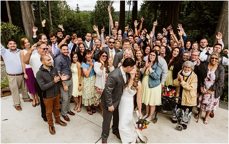 Premier Snohomish & Seattle Wedding Photographer | GSquared Weddings Photography A large group of wedding guests stands outdoors, smiling and cheering with raised hands, as a bride and groom kiss in front. The joyful scene unfolds amid trees and greenery at a beautiful Snohomish Seattle wedding.                                   | Serving Snohomish, Seattle ,                  & Beyond