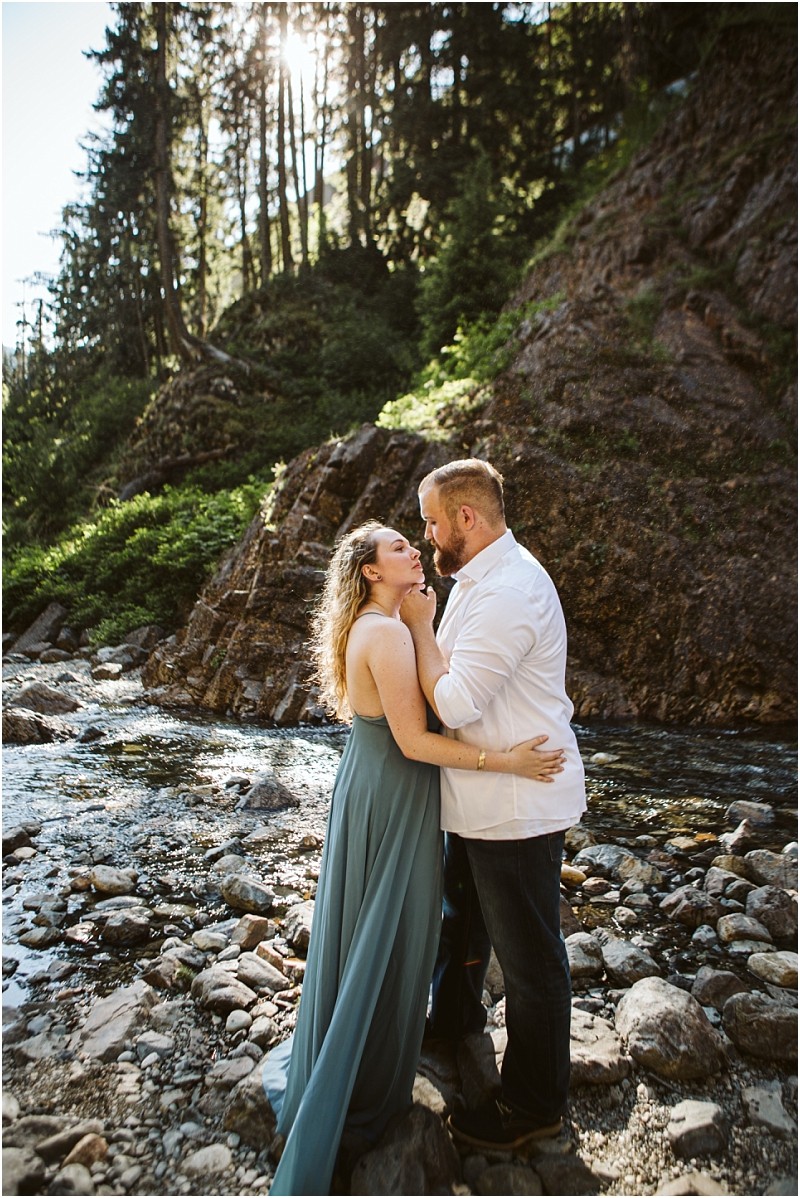 Premier snohomish & seattle wedding photographer | gsquared weddings photography a couple stands on rocky ground by a flowing stream in a forested mountainous area near snohomish The woman in a blue dress smiles up at the man in a white shirt sunlight streaming through tall trees behind themperfect for a seattle wedding engagement shoot | serving snohomish seattle                   & beyond
