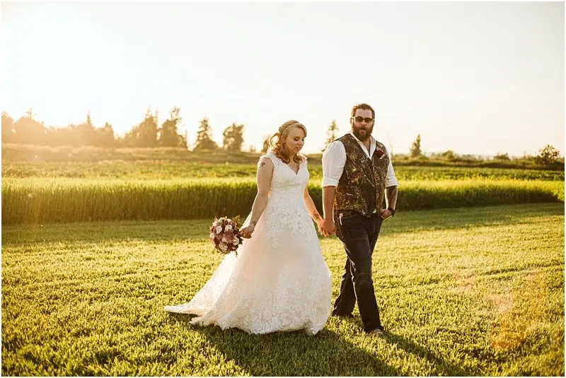 A bride in a white wedding dress holding a bouquet and a groom in a vest and jeans walk hand in hand across a grassy Snohomish field at sunset. Snohomish Wedding Photography