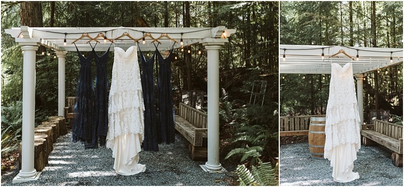 A white lace wedding dress and several navy blue bridesmaid dresses hang on wooden hangers from a white pergola in a forested snohomish outdoor setting with benches and string lights Snohomish wedding photography