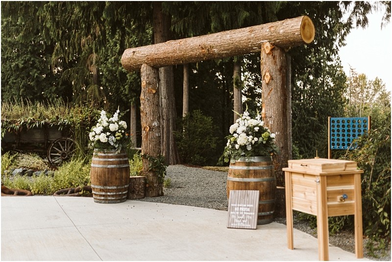 A rustic outdoor wedding altar made of large wooden logs is set on a concrete patio in snohomish flanked by barrels with white flower arrangements A wooden game table and sign stand nearby with trees and greenery filling the background Snohomish wedding photography