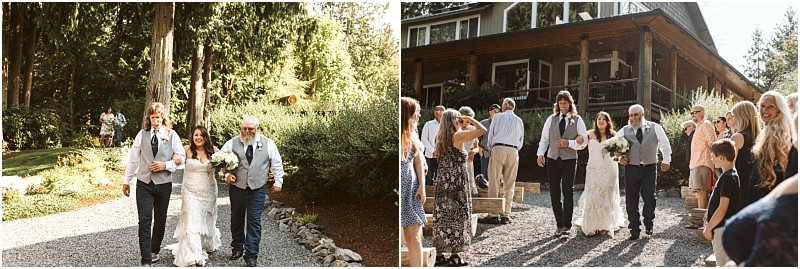 A bride in a white dress walks between two men down a gravel path outdoors in snohomish surrounded by trees and greenery with guests standing and watching near a wooden building Snohomish wedding photography