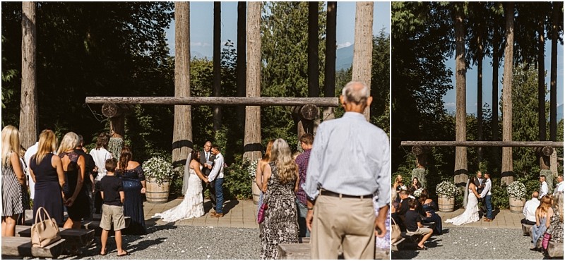A bride and groom kiss at an outdoor snohomish wedding ceremony surrounded by tall trees with guests seated and standing watching the couple Sunlight filters through the greenery Snohomish wedding photography