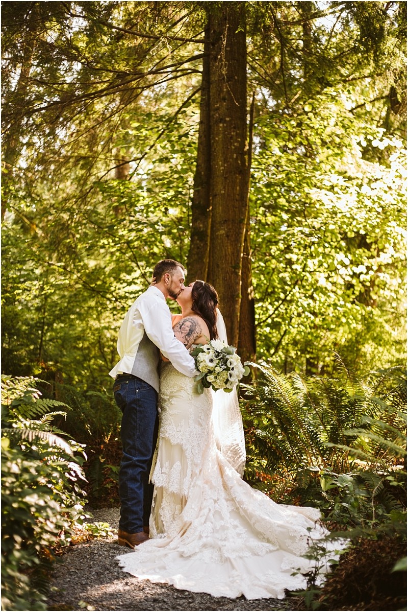 A bride in a lace gown holding a bouquet kisses a groom in a vest and jeans as they stand on a forest path near snohomish surrounded by lush greenery and sunlight filtering through the trees Snohomish wedding photography