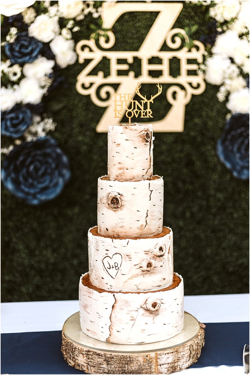 A four tiered wedding cake designed to look like birch tree logs with a heart and j+b carved into it sits on a wooden slab a gold topper reads the hunt is over perfect for a rustic snohomish celebration amid florals and greenery Snohomish wedding photography