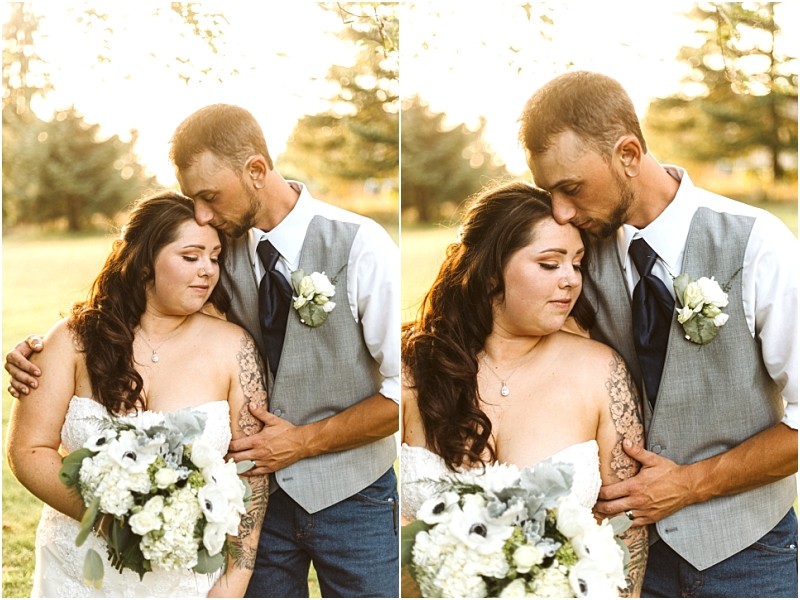 A bride holding a white bouquet stands beside a groom in a gray vest and tie He gently kisses her forehead in this romantic snohomish outdoor setting surrounded by lush greenery and sunlight Snohomish wedding photography