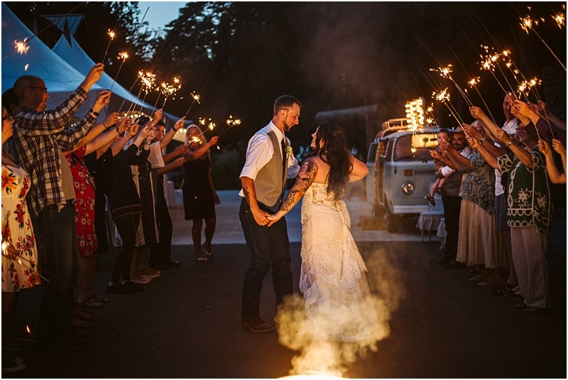 A newlywed couple holds hands and kisses while walking through a crowd of guests holding sparklers at dusk celebrating outdoors in snohomish with a decorated vintage van in the background Snohomish wedding photography